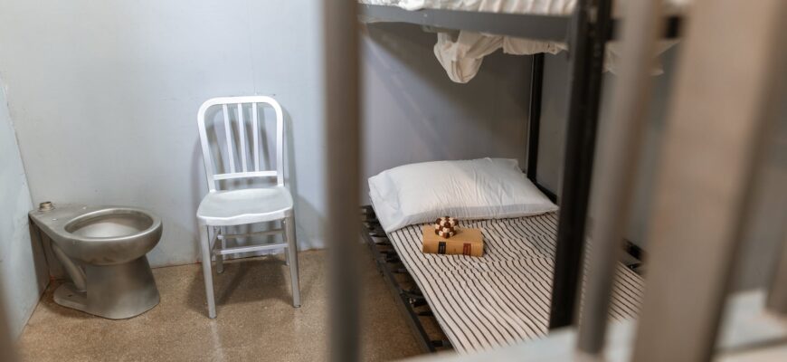 a bunk bed near a chair and toilet behind steel bars