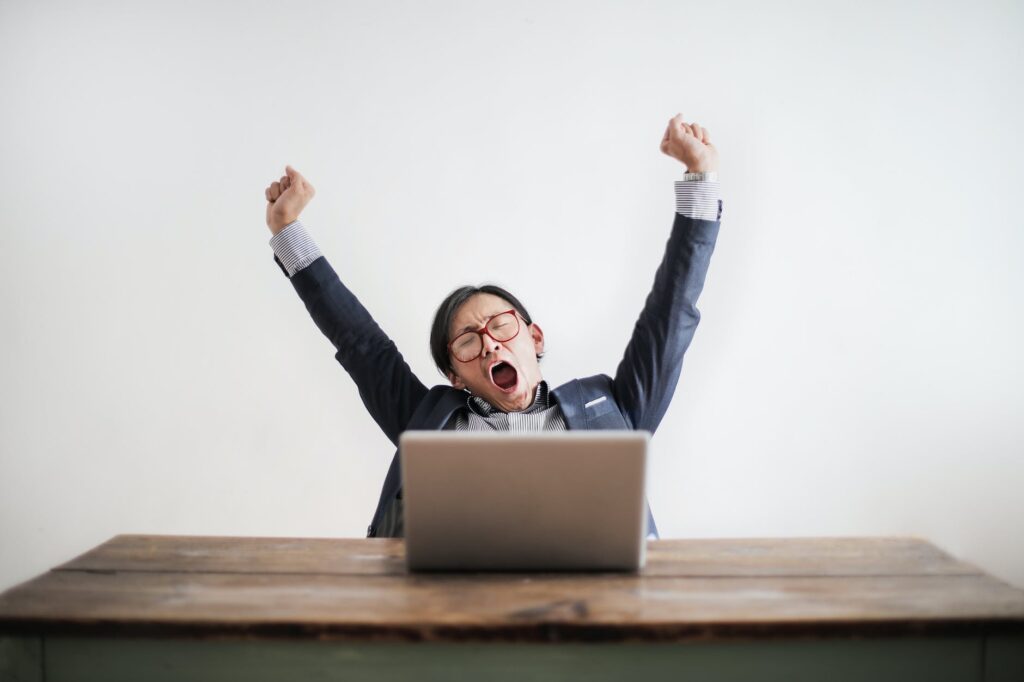 photo of yawning man with his hands up and eyes closed sitting at a table with his laptop