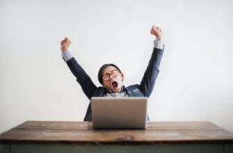 photo of yawning man with his hands up and eyes closed sitting at a table with his laptop