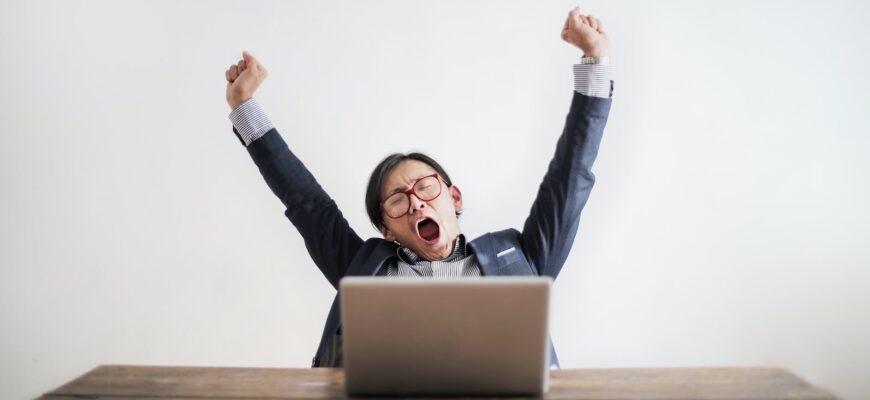 photo of yawning man with his hands up and eyes closed sitting at a table with his laptop